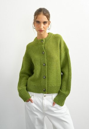 Young woman with tied-back hair wearing silver hoop earrings, a green buttoned cardigan, and white pants against a plain white background.