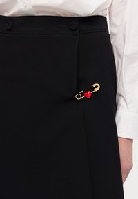 Sort nederdel lavet af glat stof har en guld sikkerhedsnål accent med en rød hjerte detalje, placeret nær lommen på venstre side.