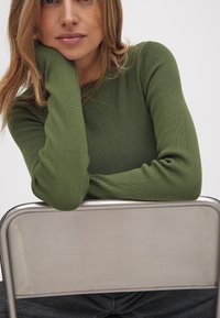 Green ribbed long-sleeve top with fitted sleeves, paired with gray denim. Silver metal chair in background, smooth texture and clean design.