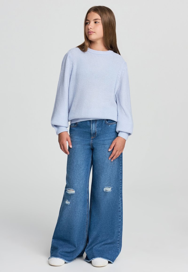 Nastolatka w jasnoniebieskim swetrze z dzianiny i szerokich, podartych dżinsach, stoi z rękami luźno opuszczonymi, patrząc w lewo na jednolitym tle.
