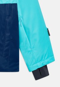 Termojacka med en färgblockerad design. Överdelen är i klar aqua, nedre delen i marinblått. Innehåller en zipficka och justerbar mudd. Slät textur.