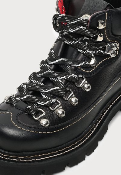 Botte de randonnée en cuir noir avec semelle épaisse, crochets métalliques pour les lacets et lacets à motif noir et blanc.