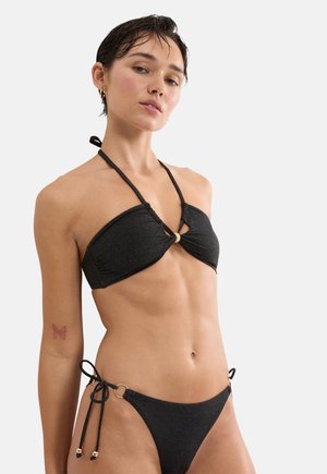 Vrouw met kort nat haar, gekleed in een zwart glinsterende bikini met een halternek top en bikini broekje met zijstrik, staand tegen een witte achtergrond.