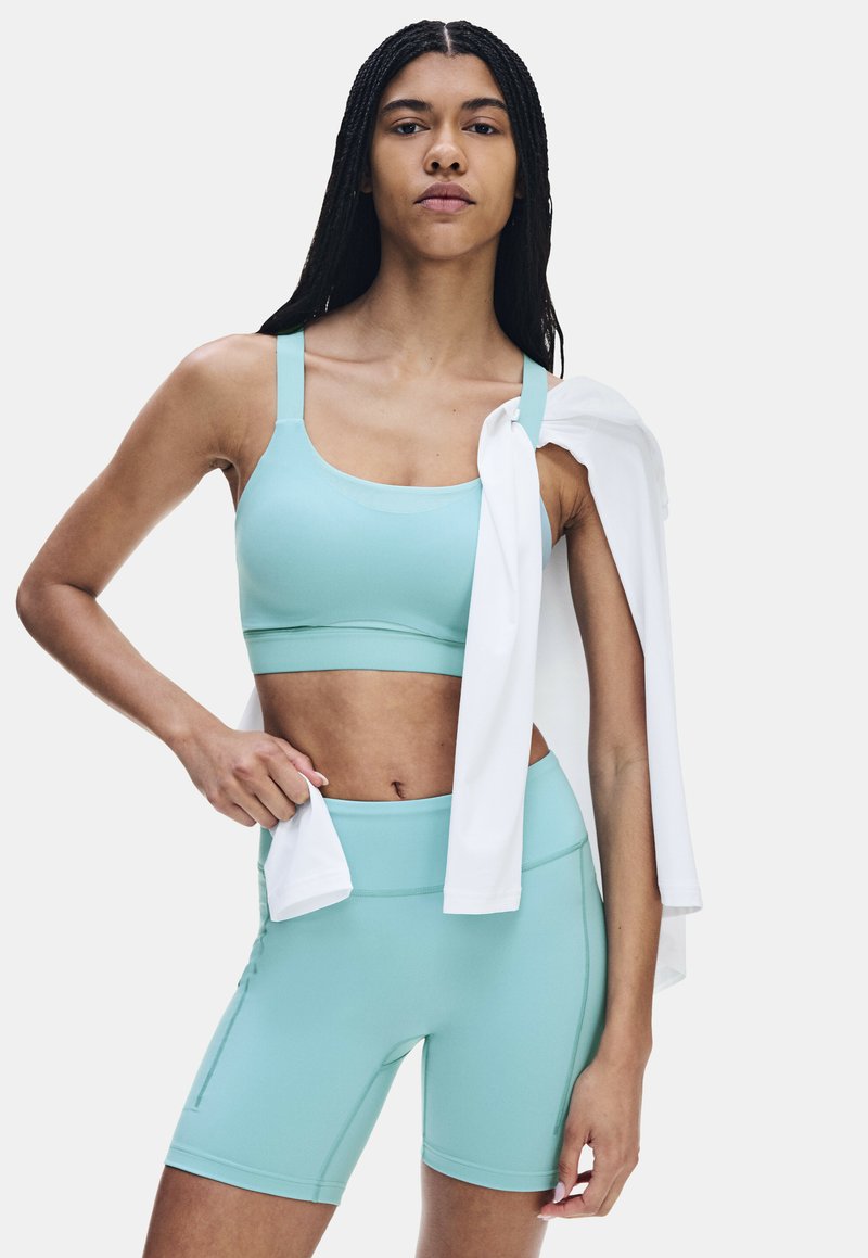 Femme vêtue d’une brassière de sport aqua et d’un short cycliste assorti, debout avec confiance, une veste blanche posée sur l’épaule.