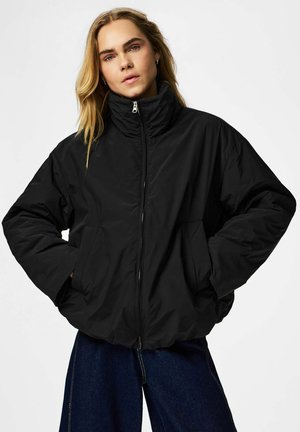 Jeune personne aux longs cheveux blonds portant une doudoune noire, les mains dans les poches, et un jean bleu foncé sur fond blanc.