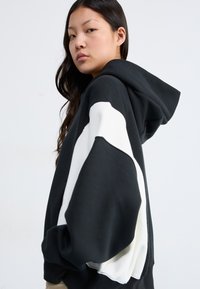 Hoodie noir avec un motif de vagues blanches sur les manches, doté d'une capuche et d'une coupe ample. Le tissu doux et les bord-côtes côtelés rehaussent la texture.