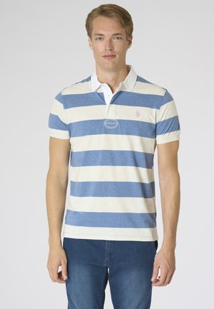 Giovane uomo in piedi, indossa una polo a righe blu e bianche a maniche corte e jeans blu su uno sfondo semplice.
