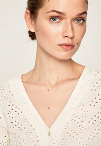 Blusa bianca con tagli a occhio di pernice, colletto a V profondo e chiusura con bottoni. Tessuto testurizzato con un design delicato e arioso.
