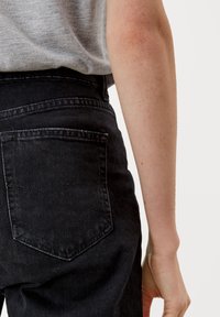 Czarne jeansy z denimu mają wysoki stan, przeszycia oraz jednolitą tylną kieszeń. Tkanina wydaje się miękka z delikatną fakturą.