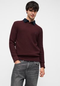 Pull en maille bordeaux avec finition texturée et col à revers bleu marine, associé à un jean gris. Logo discret sur la poitrine.