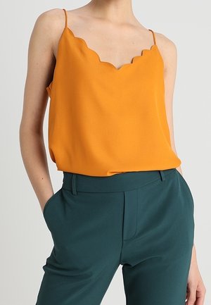 Kobieta w pomarańczowym topie bez rękawów z falowanym brzegiem, wsadzonym w ciemnozielone spodnie z wysokim stanem, z rękami w kieszeniach.
