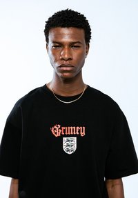 Camiseta negra de algodón con texto "Grimey" en rojo y un gráfico de escudo con un diseño estilizado. Corte estándar con mangas cortas.
