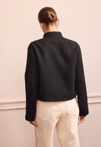 Giacca corta nera con texture, collo alto e maniche lunghe, abbinata a pantaloni chiari a vita alta.