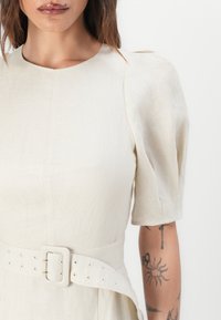 Vaaleanbeige pellavamekko, jossa on lyhyet puffihihat ja strukturoitu pääntie. Mekossa on leveä vyö neliösoljella ja näkyviä napinreikiä.