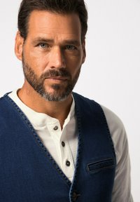 Jeansväst med en vit henley-tröja; har blått tyg, en ficka och kontrasterande sömnad på västen. Neutral bakgrund.