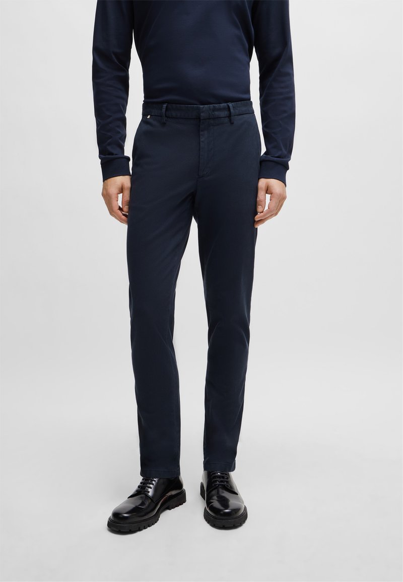 Navy blue trousers made of cotton-blend fabric, slim fit design, featuring a flat front, belt loops, and a smooth finish. Paired with black shoes.