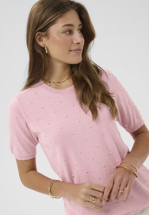 Ung kvinde med langt brunt hår iført en pink tekstureret top med korte ærmer og guldsmykker, der kigger mod højre mod en ensfarvet baggrund.