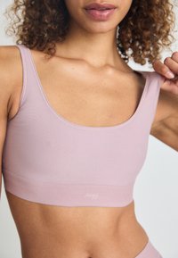 Top sportivo rosa chiaro con scollatura ampia e spalline spesse. Tessuto liscio con una leggerissima texture, con un piccolo logo nella parte inferiore.