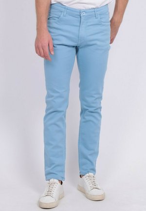 Homme portant un pantalon slim bleu clair et des baskets blanches, debout devant un fond clair uni.