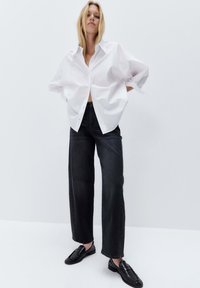 Chemise blanche surdimensionnée à boutons, jeans larges noirs, et mocassins noirs. Tissu lisse et coupe ample ; style décontracté et contemporain.