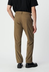 Braune maßgeschneiderte Hosen aus glattem Stoff, mit einer taillierten Passform, Gesäßtaschen und kombiniert mit schwarzen Schuhen, von der Seite sichtbar.