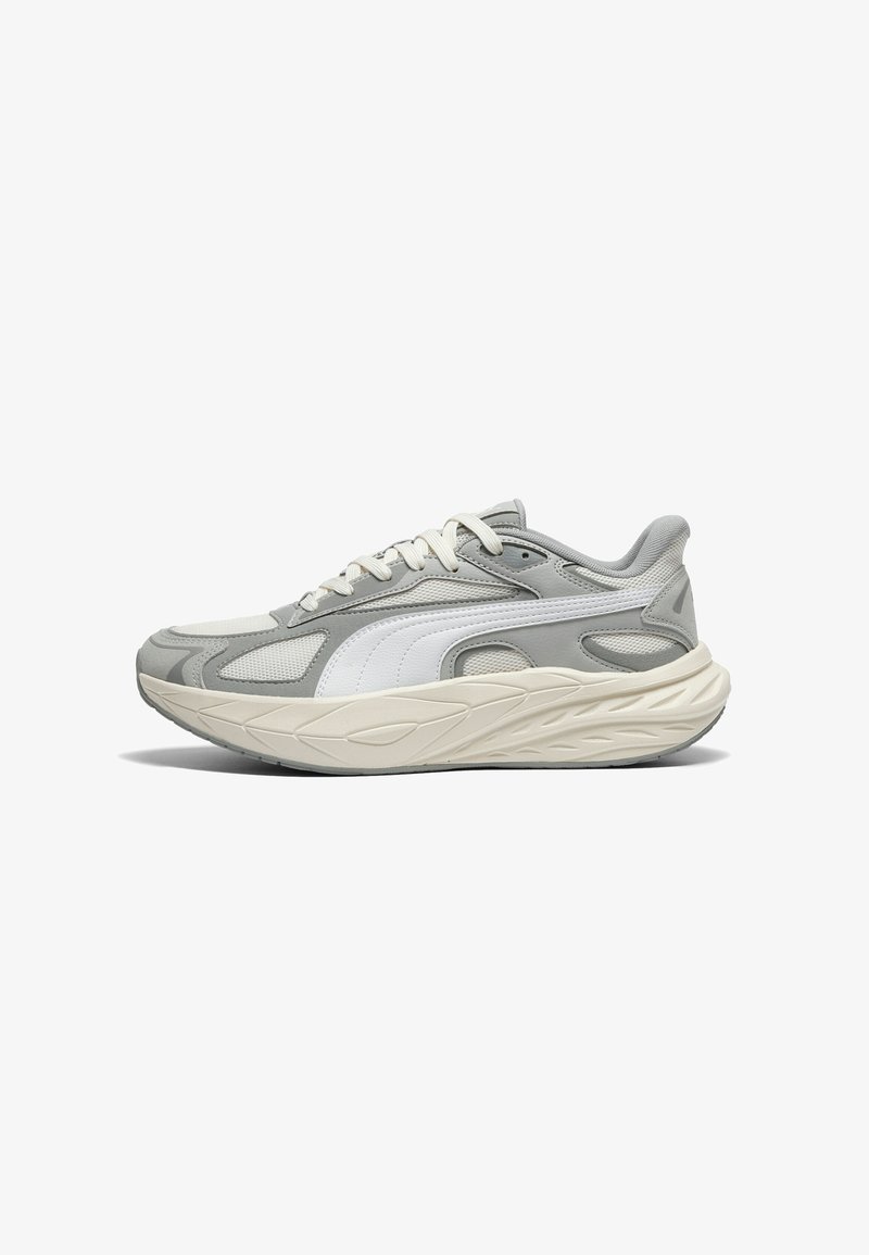 Chaussure de sport grise en mesh et matériau synthétique. Présente des accents texturés, une semelle beige clair et une fermeture à lacets. Logo latéral notable.