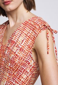 Väri-richön ilman hihoja mekko, jossa on geometrinen kuvio punaisessa, oranssissa ja kermassa. Mukana solmimisnauhat olkapäillä ja syvä V-kaula-aukko.