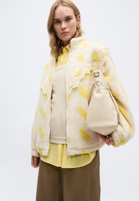 Νέα γυναίκα που φοράει κίτρινο πουκάμισο, εκρού φλις μπουφάν με κίτρινα σχέδια και δαντέλα στο τελείωμα, καφέ παντελόνια, κρατώντας μπεζ τσάντα στον δεξιό ώμο.