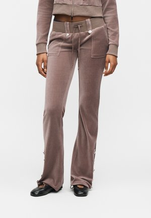 Persona che indossa pantaloni svasati in velluto malva con tasche anteriori, vita con coulisse, dettagli con bottoni sui lati, abbinati a ballerine nere.