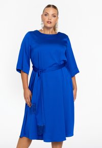 Blauwe satijnen jurk met korte mouwen, tailleband en zij-tassel detail. Gladde textuur, knielengte en een ronde halslijn.