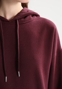 Sudadera burdeos hecha de tejido suave, con capucha clásica, cordones con puntas plateadas y un ajuste holgado. Textura lisa, sin patrones.