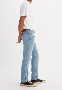 Homme portant un jean bleu clair, un t-shirt blanc et des baskets noires, debout de profil les mains jointes derrière le dos sur fond blanc.