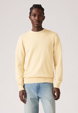 Mężczyzna z zaplecionymi włosami, ubrany w pastelowy żółty sweter z okrągłym dekoltem i jasnoniebieskie dżinsy, stojący na tle prostego, jasnego tła.