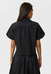Zwarte cropped shirt met korte pofmouwen en een puntige kraag. Achterkant heeft een centrale naad en een gladde, licht gestructureerde stof.