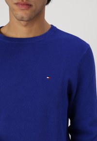Pull en tricot bleu avec un tissu texturé, col rond, et un petit logo brodé en rouge, blanc et bleu sur la poitrine.