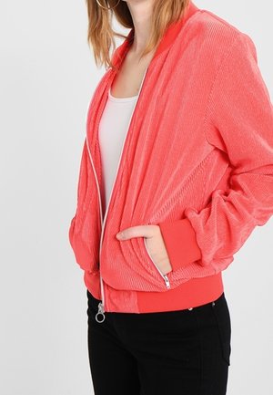 Giovane donna che indossa una giacca corallo a coste con zip sopra una canottiera bianca e pantaloni neri, con una mano nella tasca della giacca.