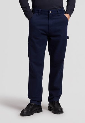 Homme portant un pantalon décontracté bleu marine et des chaussures noires à lacets, les mains dans les poches, debout devant un fond clair uni.