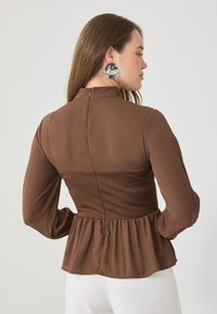 Kobieta z długimi brązowymi włosami, ubraną w dopasowaną brązową bluzkę z długimi rękawami i peplum, zestawioną z białymi spodniami, zwróconą bokiem.