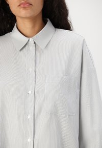 Camicia a righe grigia e bianca, con colletto, maniche lunghe e taschino sul petto. Il materiale sembra leggero e liscio.