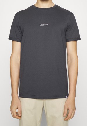 Mężczyzna noszący ciemnoszary T-shirt z krótkim rękawem z napisem „LES DEUX” w białym kolorze na środku klatki piersiowej, w połączeniu z jasnobeżowymi spodniami.