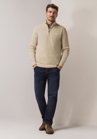 Beige ribgebreide trui met ritskraag, gecombineerd met donkerblauwe jeans en bruine suède schoenen. Model staat ontspannen met handen in zakken tegen een neutrale achtergrond.