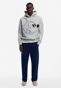 Sudadera con capucha gris con diseño azul de Mickey Mouse, corte relajado, combinada con pantalones azul marino y zapatos de color claro. Textura de tela suave.