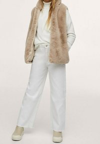 Faux fur gilet color crema sopra un maglione bianco e jeans bianchi a vita alta. L'outfit presenta una vestibilità comoda e scarpe beige slip-on.