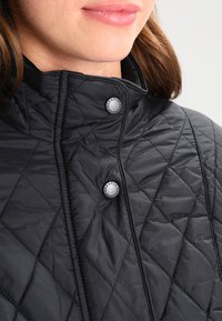 Casaco preto acolchoado com gola alta, fechos de pressão e uma textura lisa e leve. Costura em padrão de losangos notável.