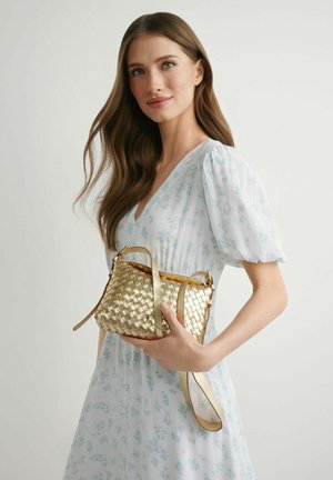 Kobieta z długimi brązowymi włosami w białej sukience w kwiaty, trzymająca plecioną złotą torebkę na jednolitym tle.