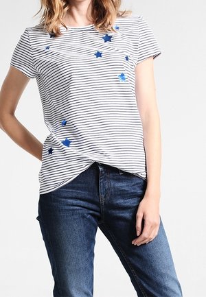Frau trägt ein weißes und marineblau gestreiftes T-Shirt mit kurzen Ärmeln und blauen Stern-Applikationen, kombiniert mit blauen Jeans.