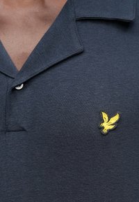 Tengerészkék póló puha anyagból, hajtókás gallérral, bal mellrészen sárga hímzett sas díszítéssel és gombos részlettel.