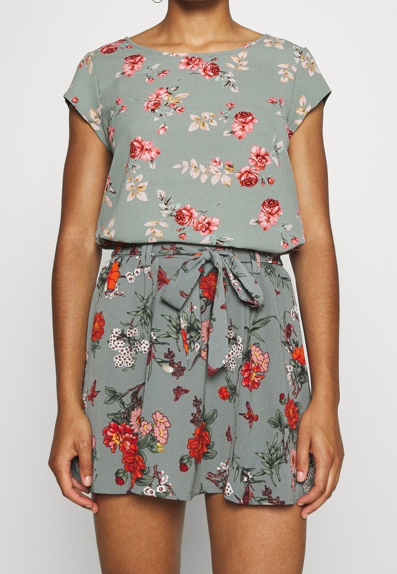 Blumiger Jumpsuit aus grünem Stoff mit kurzen Ärmeln, einem Bindegürtel in der Taille und verschiedenen Blumenmustern in Rot, Pink und Weiß.