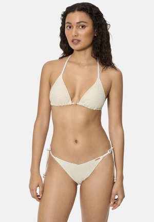 Mujer con cabello rizado y oscuro, vistiendo un bikini texturizado de color blanco roto con tirantes de lazo, de pie contra un fondo claro y sencillo.