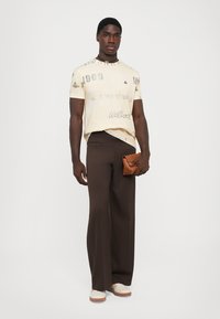Tricou grafic crem cu text imprimat, pantaloni largi maro și o poșetă bej. Modelul poartă adidași deschiși și un colier din mărgele.